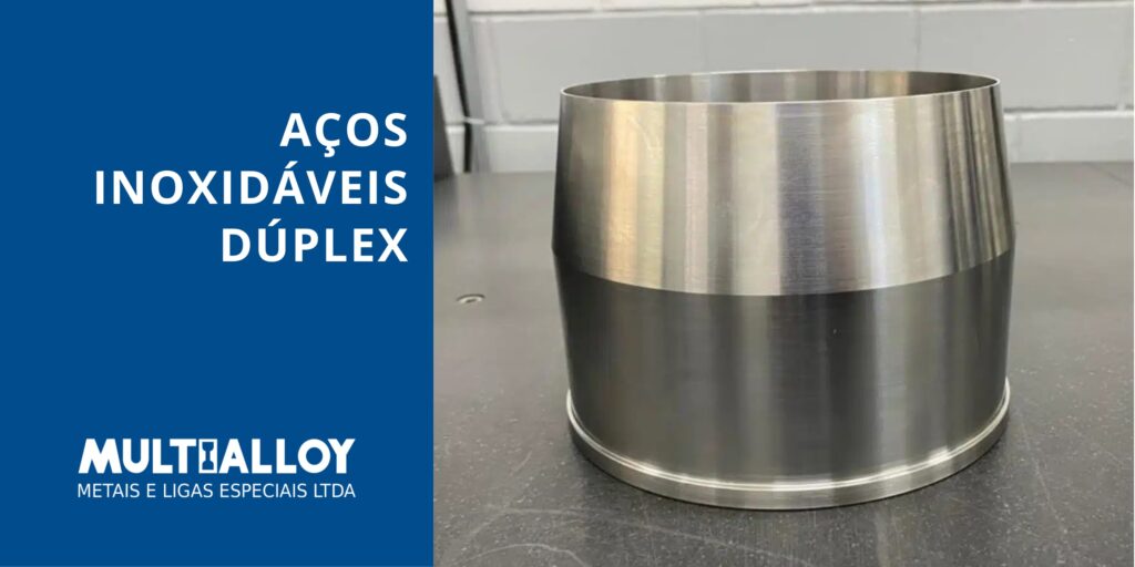 Peça metálica cilíndrica com acabamento usinado liso, apresentando um chanfro preciso na borda. O fundo é neutro e o texto à esquerda diz "Aços Inoxidáveis Dúplex"
